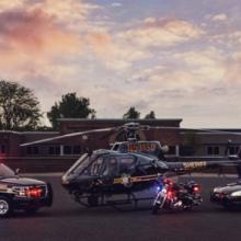 An Oakland County Sheriff’s Office SUV, helicopter, motorcycle, and cruiser with emergency lights activated sit outside at sunset