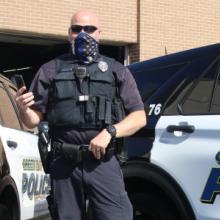 A police officer wearing a mask and a vest stands in front of a Green Bay Police vehicle